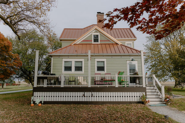 Residential house with green siding and a metal roof, featuring a large front porch and outdoor seating, surrounded by trees and autumn foliage, showcasing Knapp Builders' exterior design expertise.