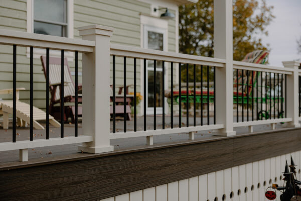 Exterior view of a residential deck featuring a white railing with black spindles, showcasing a wooden floor and a green house in the background, with outdoor seating visible.
